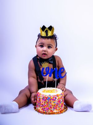 Baby with crown poses with first birthday cake decorated with sprinkles and blue 'One' topper.