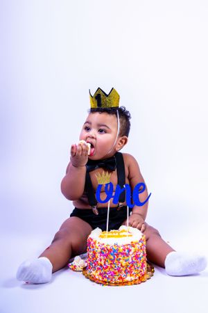 Baby in black outfit and crown gestures while sitting next to decorated birthday cake.