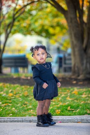 A toddler in navy blue dress stands on path surrounded by fall foliage.