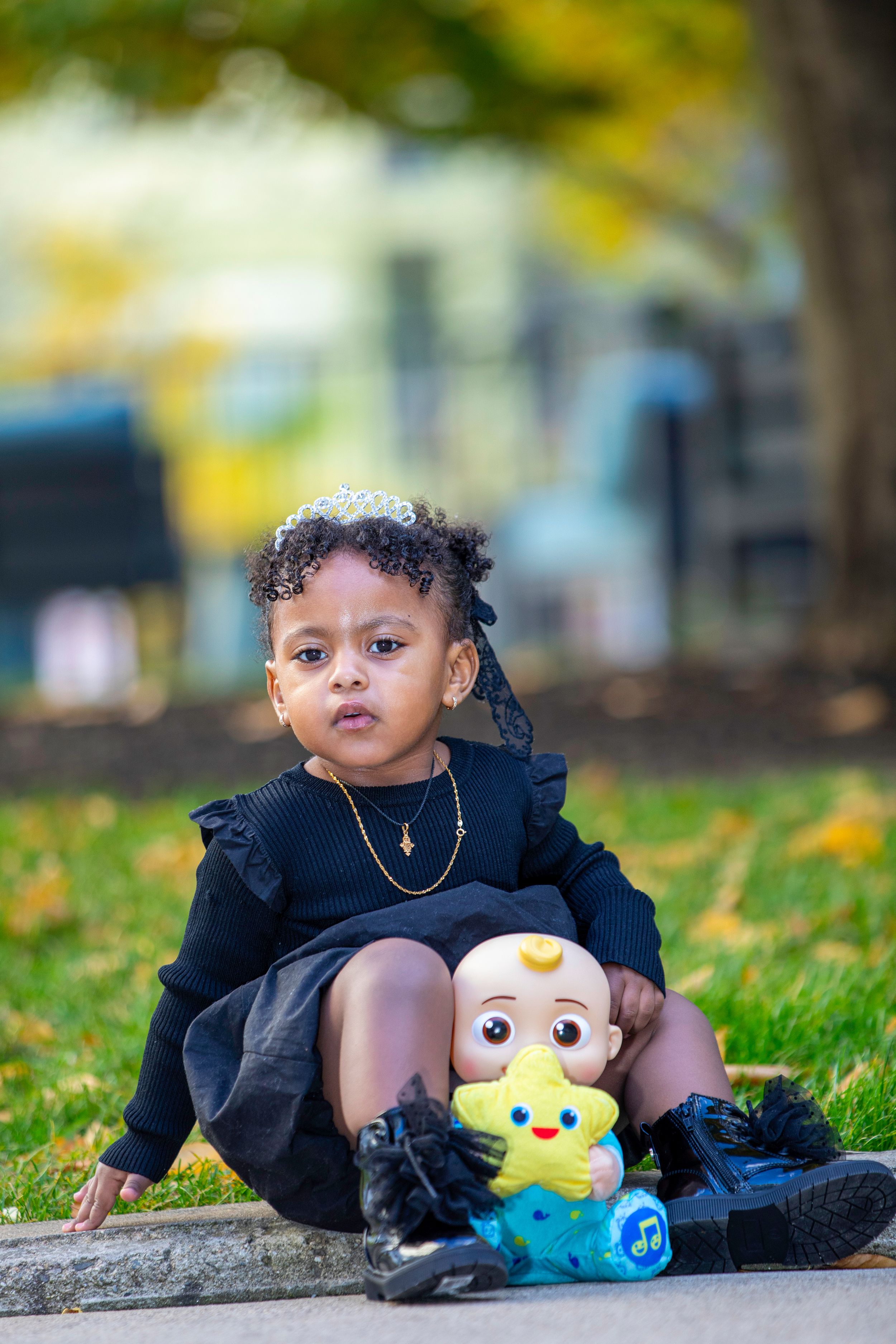 A toddler in navy dress sits with Minion toy amid autumn leaves.