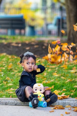 A toddler in navy dress watches falling autumn leaves with Minion toy nearby.