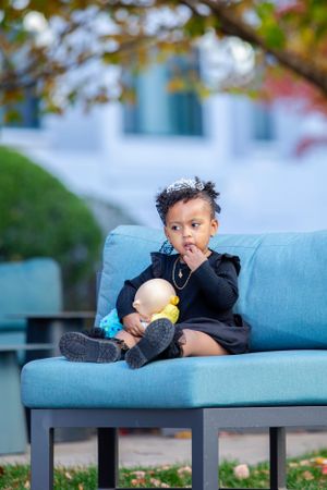 A small child in a black outfit sits thoughtfully on a bright blue outdoor couch.