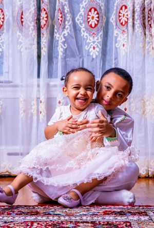 Two children in dressy white outfits sitting together on patterned carpet.