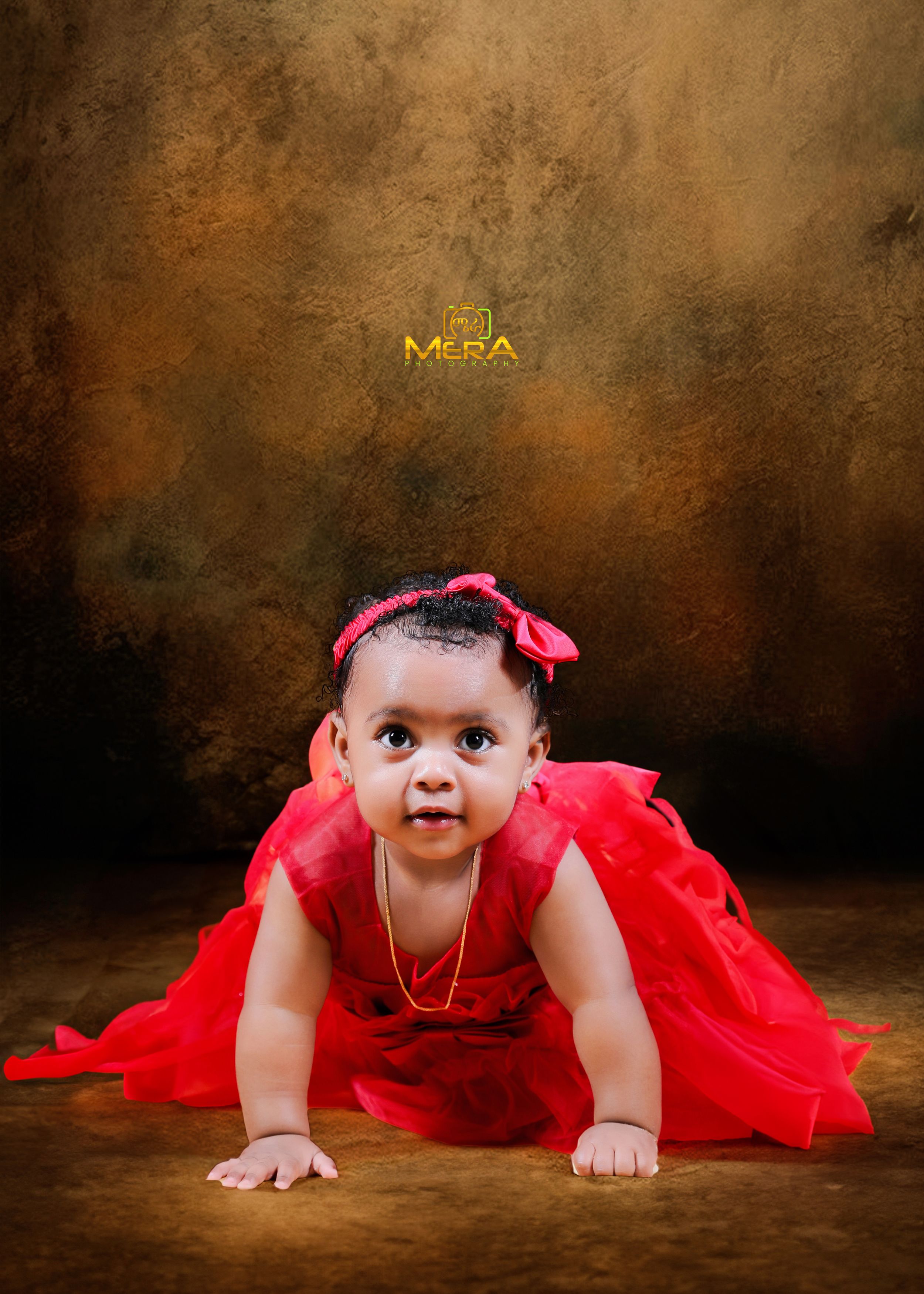 Baby in red dress crawling on brown backdrop during professional photo shoot.