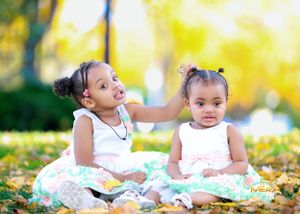 Two small children in dresses sitting together on autumn leaves.