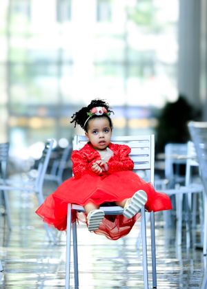 Small child wearing a red formal dress sitting on a chair in an indoor venue.