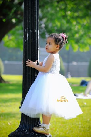 Child in white tulle dress holding onto a black lamp post in a park.