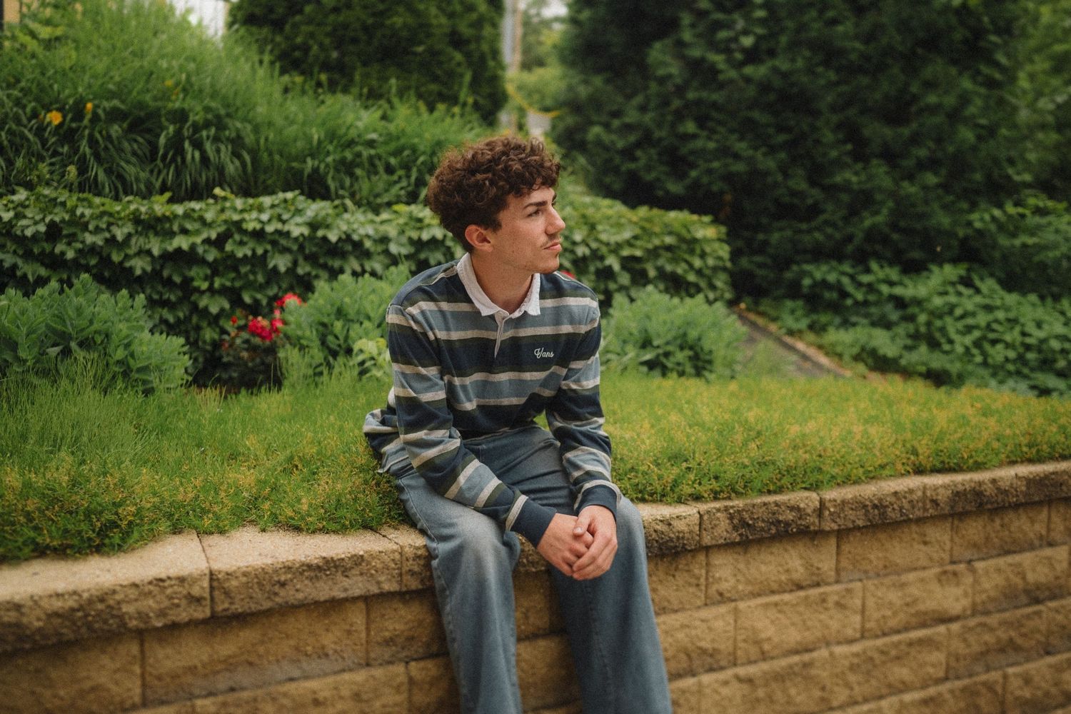 A young person in a striped sweater sits thoughtfully on a stone wall surrounded by lush garden landscaping.