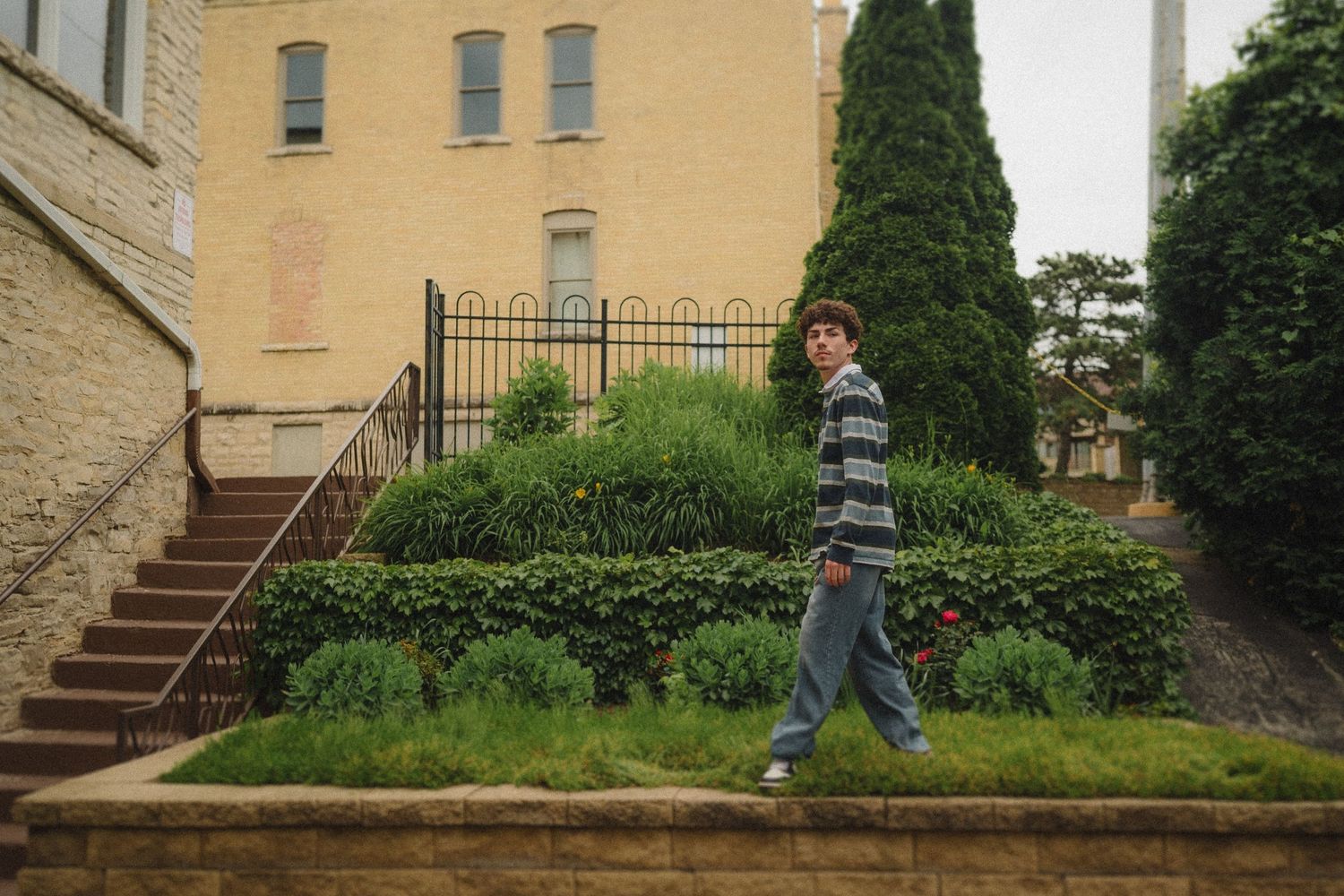 Person in striped sweater walking past historic building with landscaped garden and stone staircase.