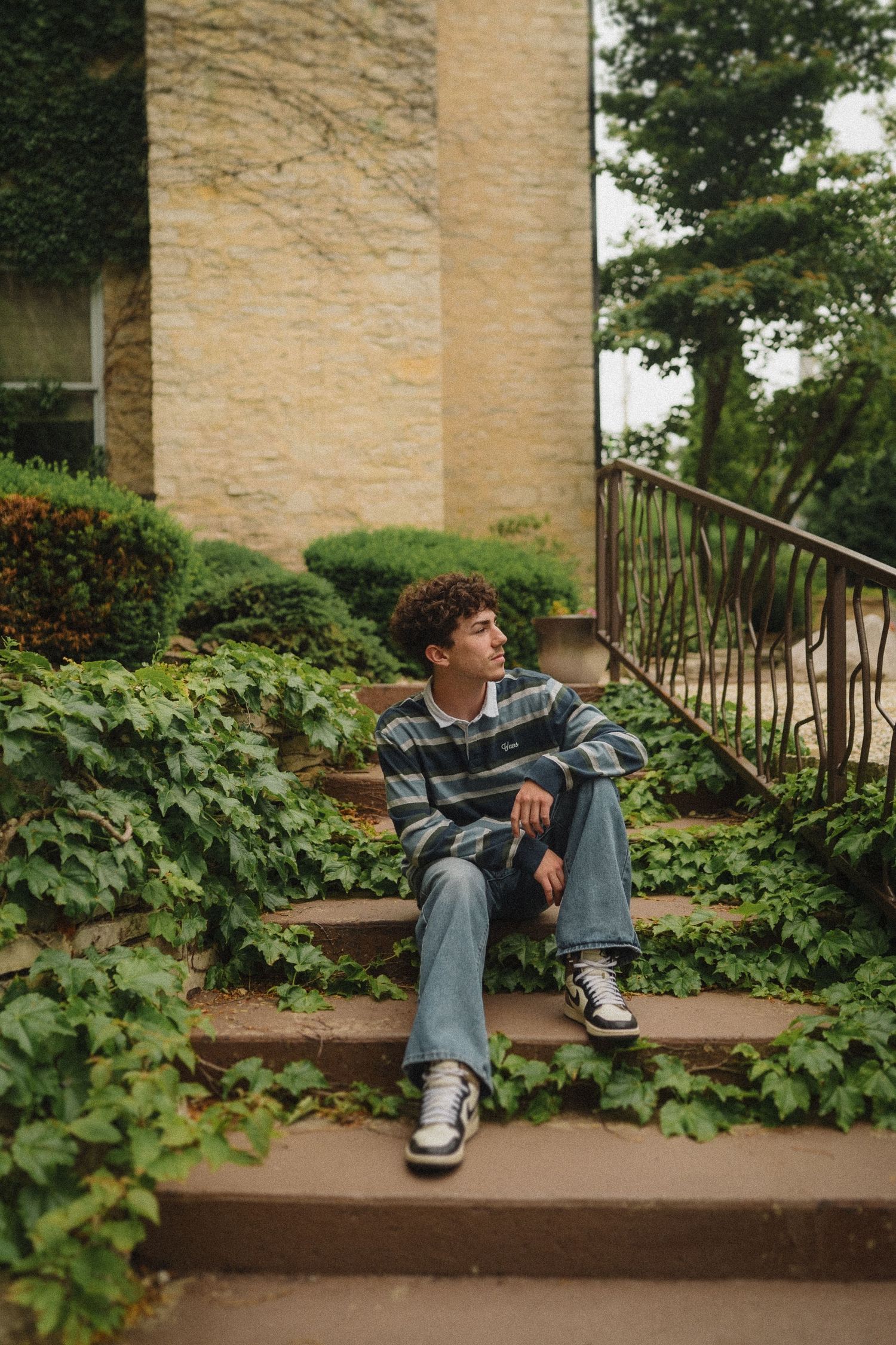 A person in a striped sweater sits casually on outdoor steps surrounded by lush ivy and stone columns.