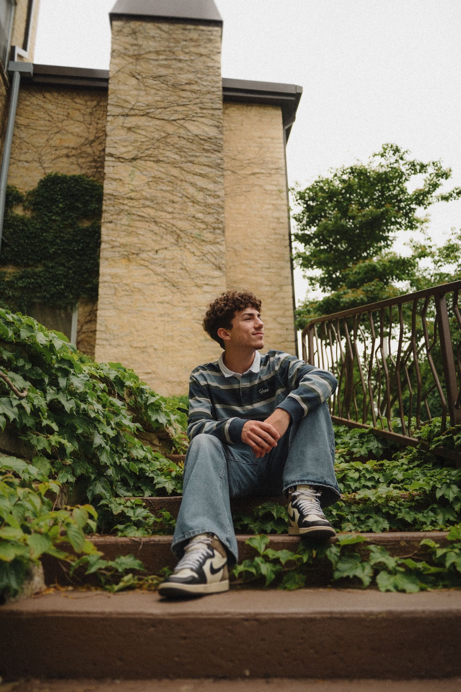 Young person in striped sweater sitting casually on stone steps surrounded by ivy and a cream-colored building wall.