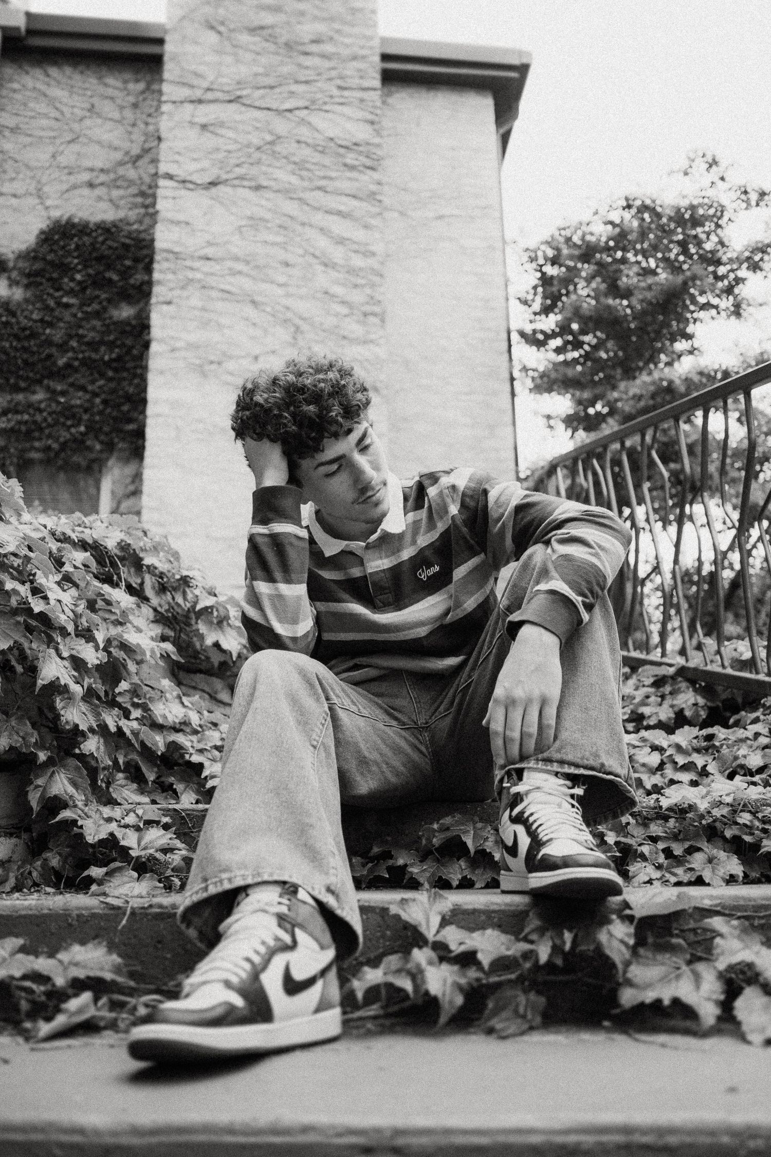 A black and white portrait of someone resting on ivy-covered steps wearing casual streetwear.