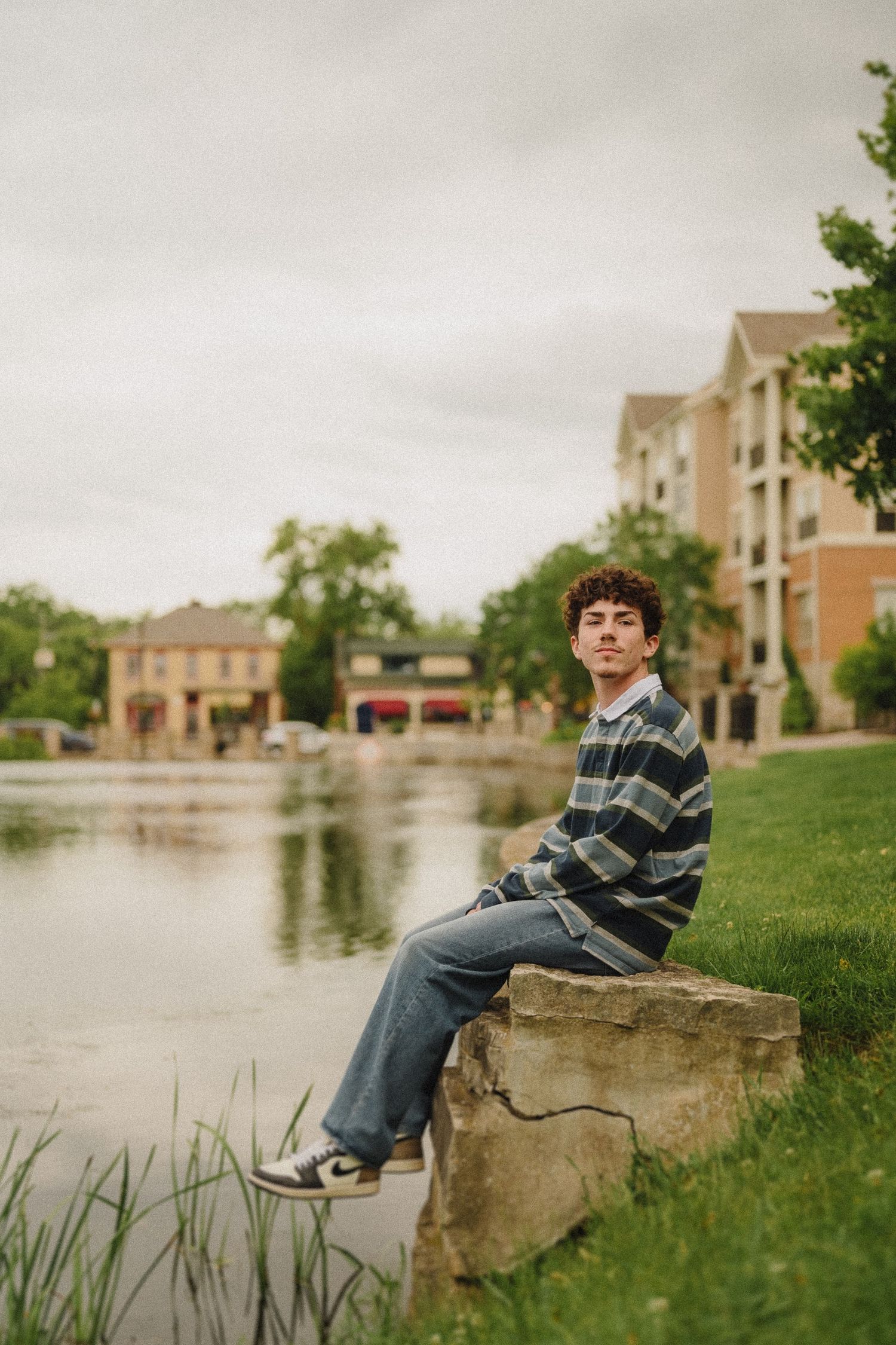 A person in a striped sweater sits contemplatively on a stone by a lake with apartment buildings in the background.