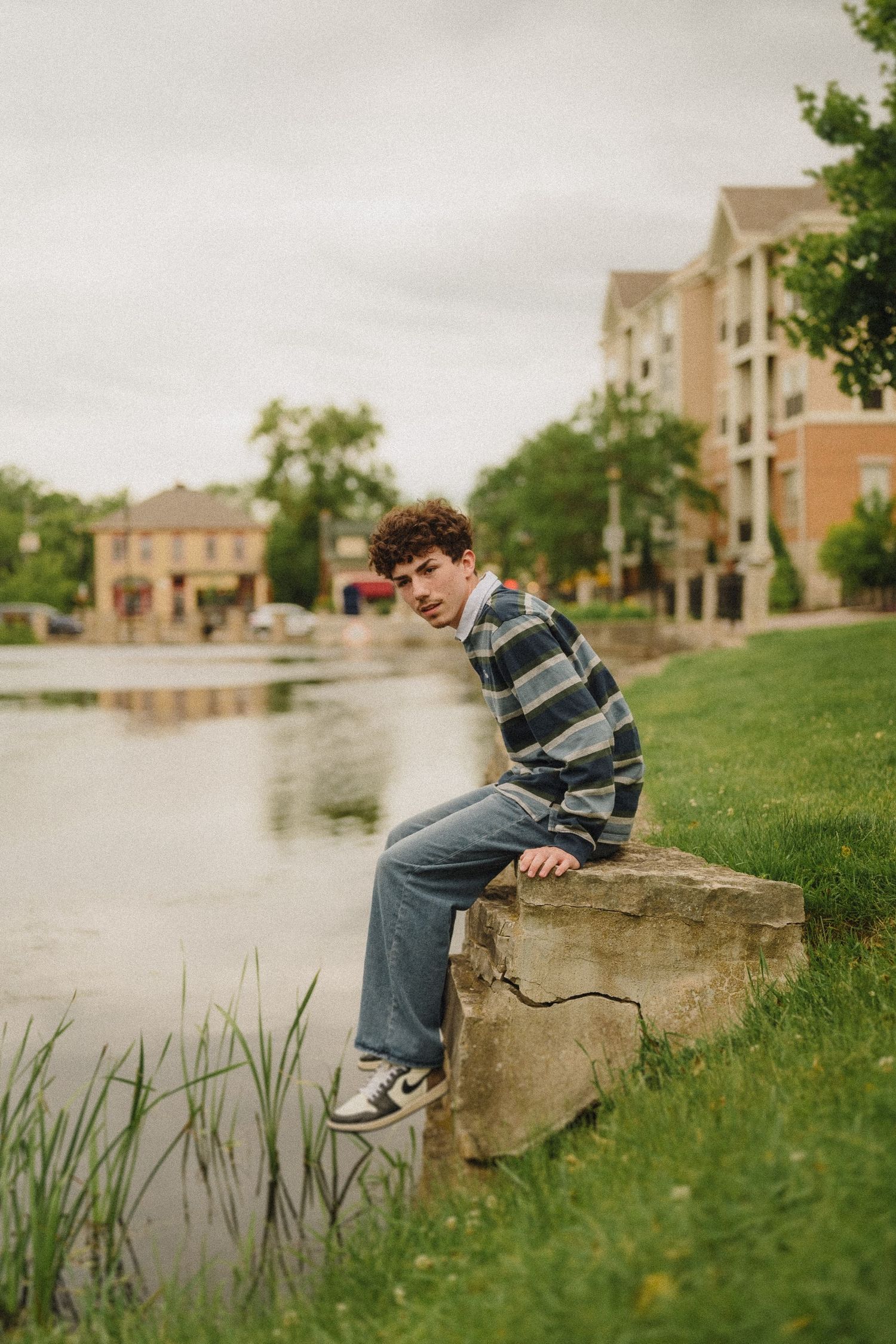 Person in striped sweater sits on a rock by a pond with apartment buildings in the background on an overcast day.