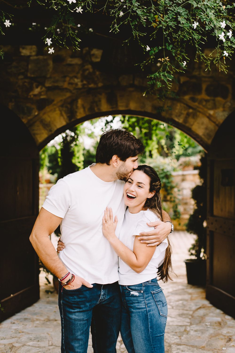Happy couple in white t-shirts embracing and laughing under a stone archway with flowers in a garden.