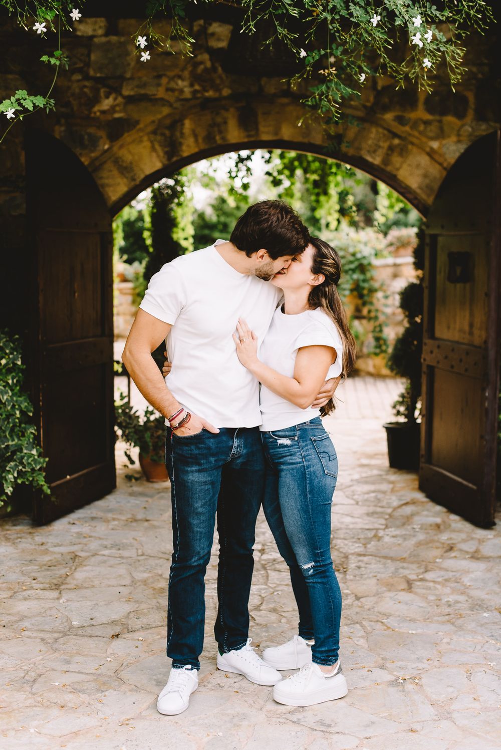 Couple kissing in front of a stone archway with lush greenery, both wearing white shirts and jeans.