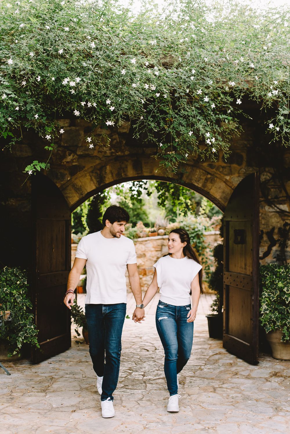 Couple holding hands walking through a stone archway covered in green vines and white flowers.