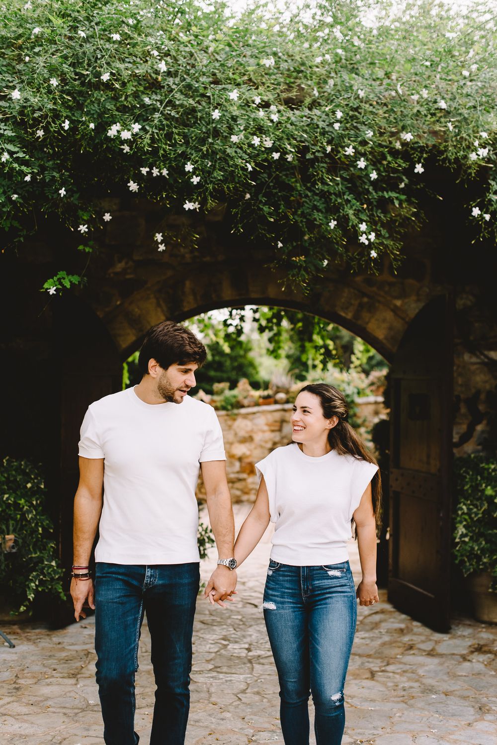 Couple holding hands and smiling while walking under a stone arch covered in white flowers.