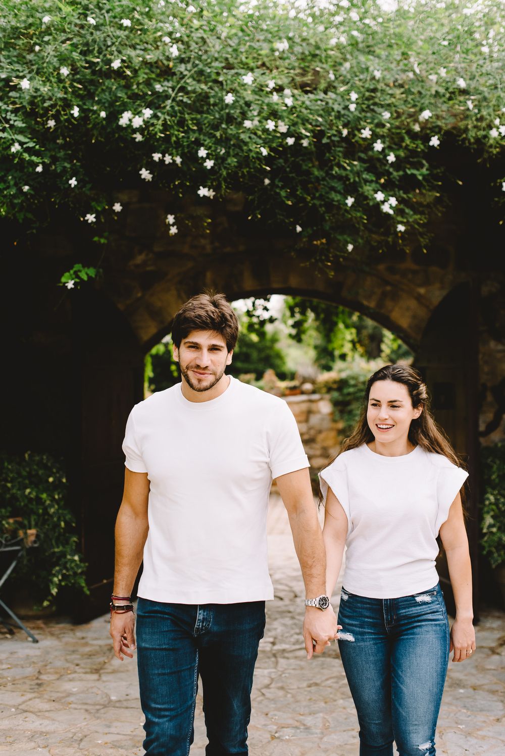 Happy couple in matching white t-shirts and jeans holding hands walking under a floral stone arch outdoors.