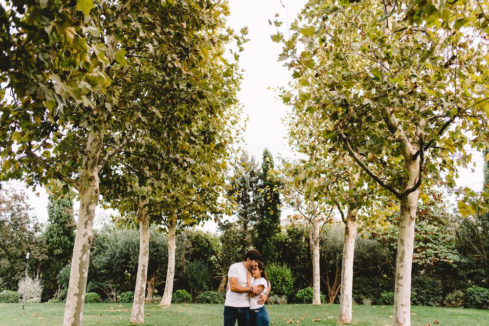 Couple embracing romantically among tall trees in a lush green park during an outdoor engagement photo session.