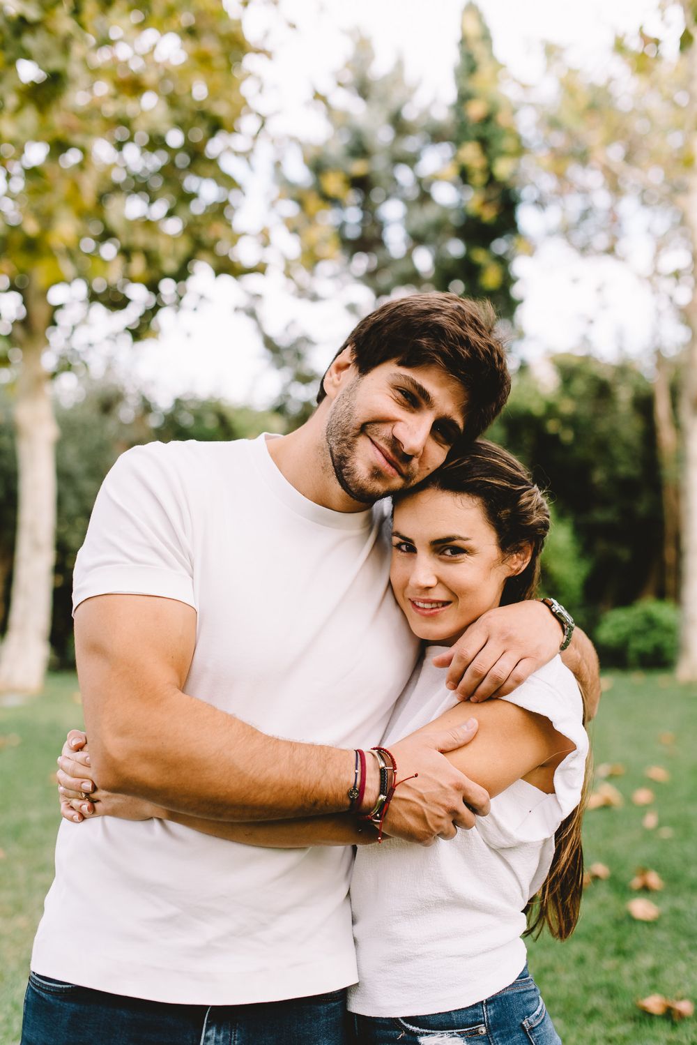 Happy couple in white t-shirts embracing in a green park, smiling warmly at the camera on a sunny day.