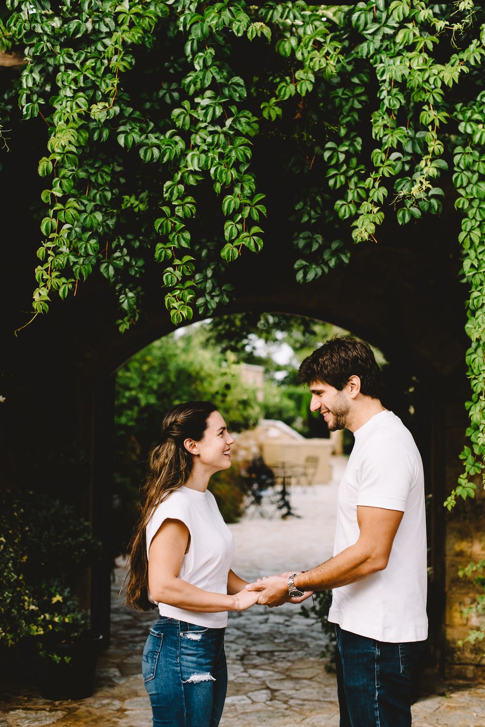Happy couple holding hands and smiling under a lush green vine archway in a romantic outdoor setting.