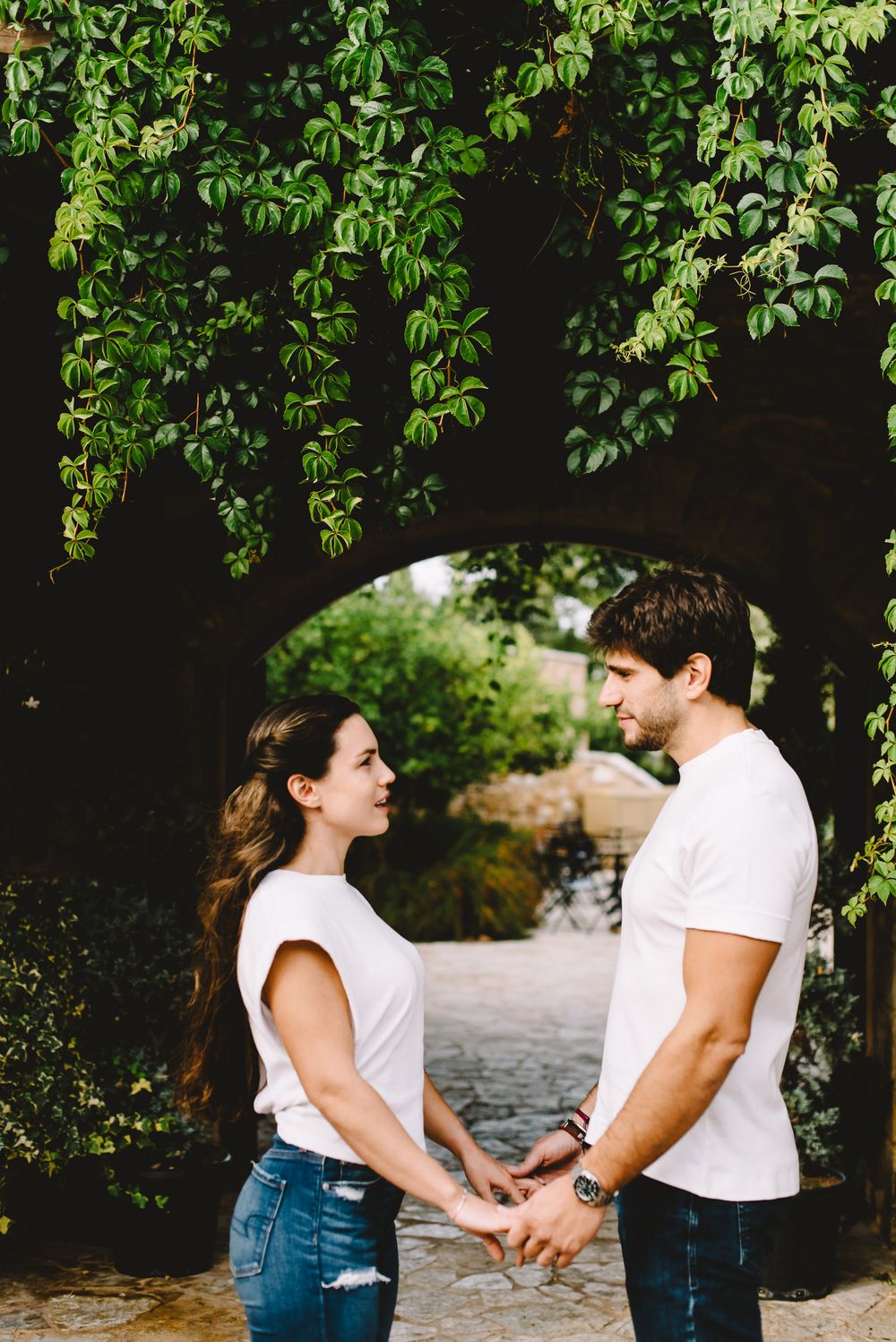 Romantic couple holding hands and gazing at each other under a lush green vine archway in a garden setting.