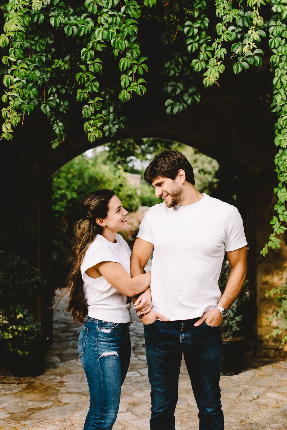 Happy young couple in matching white tees and jeans holding hands under a lush green garden archway.
