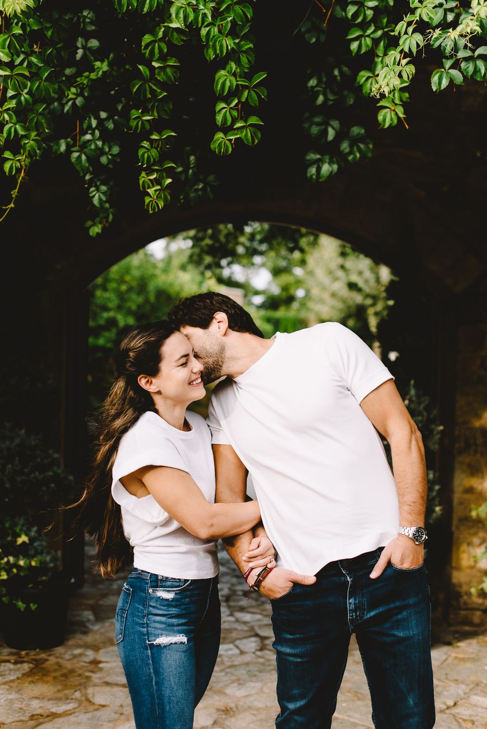 Couple in matching white tees and jeans sharing a romantic moment under a vine-covered stone archway outdoors.