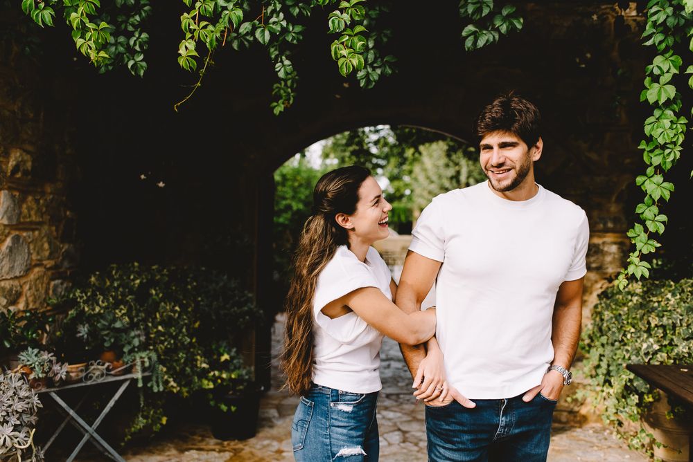 Happy young couple walking hand in hand through a vine-covered stone archway in a lush garden setting.