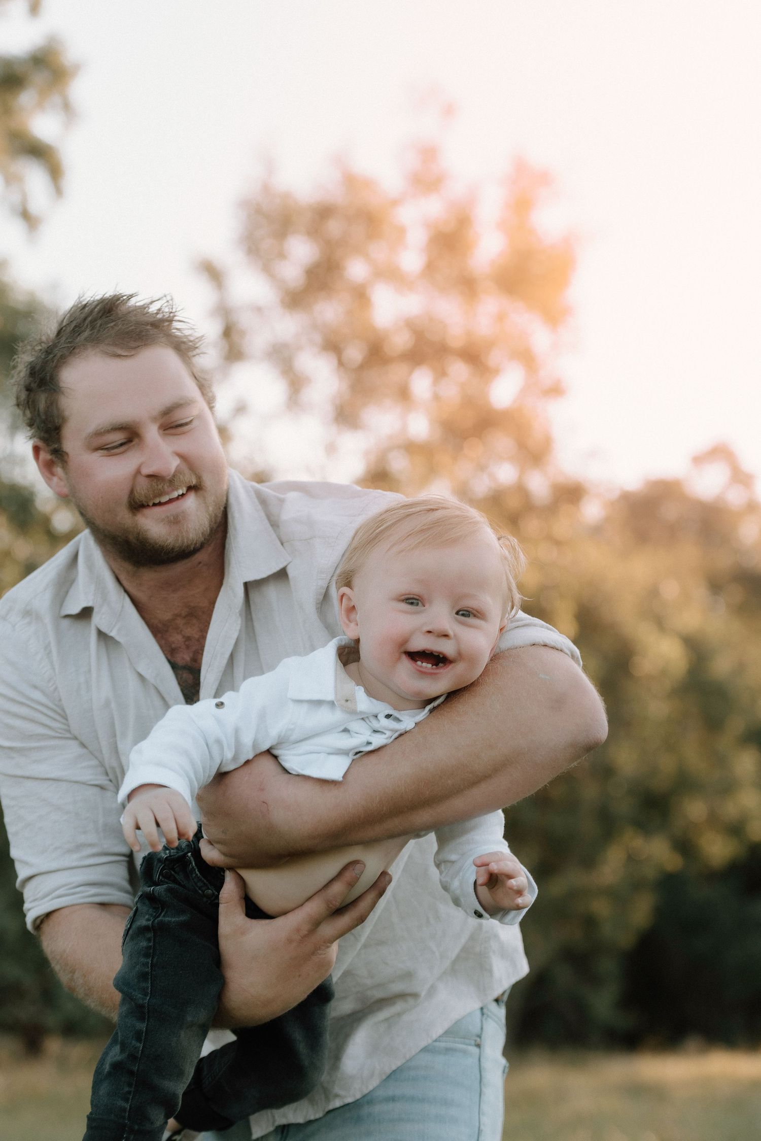 A smiling parent wearing a light shirt holds and plays with a laughing baby outdoors during golden hour. Father and Son. Traralgon, Victoria