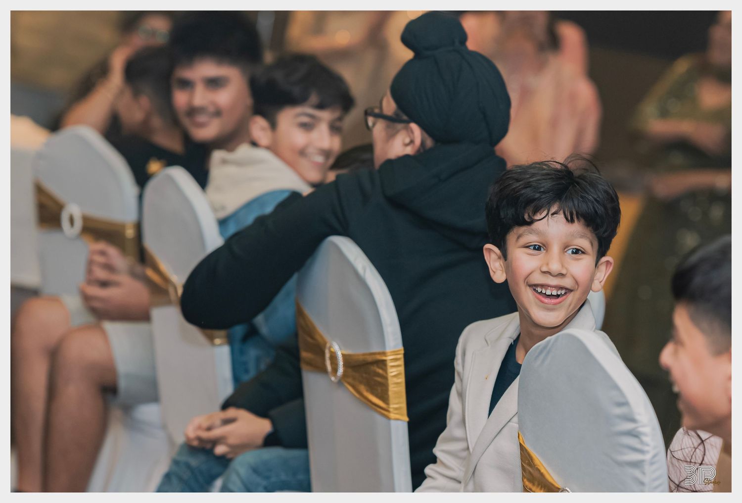 Young guests at a wedding reception smile while sitting in decorated chairs.