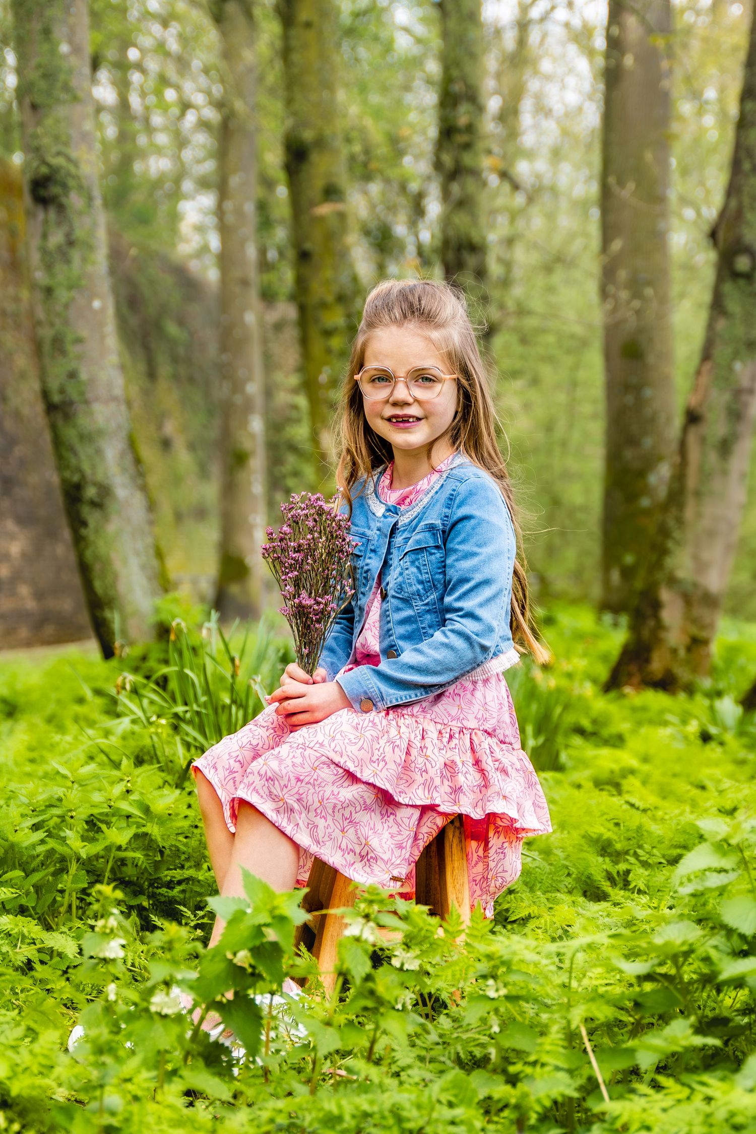 Child in blue jacket holding dried flowers while sitting in lush green forest setting.