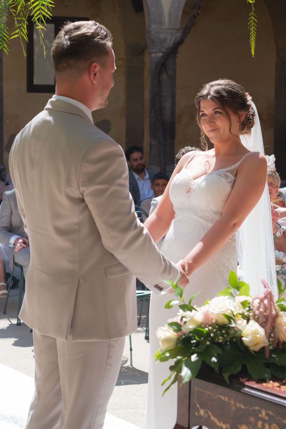 A bride and groom during their wedding ceremony holding hands. The bride is looking at the groom with the biggest smile.