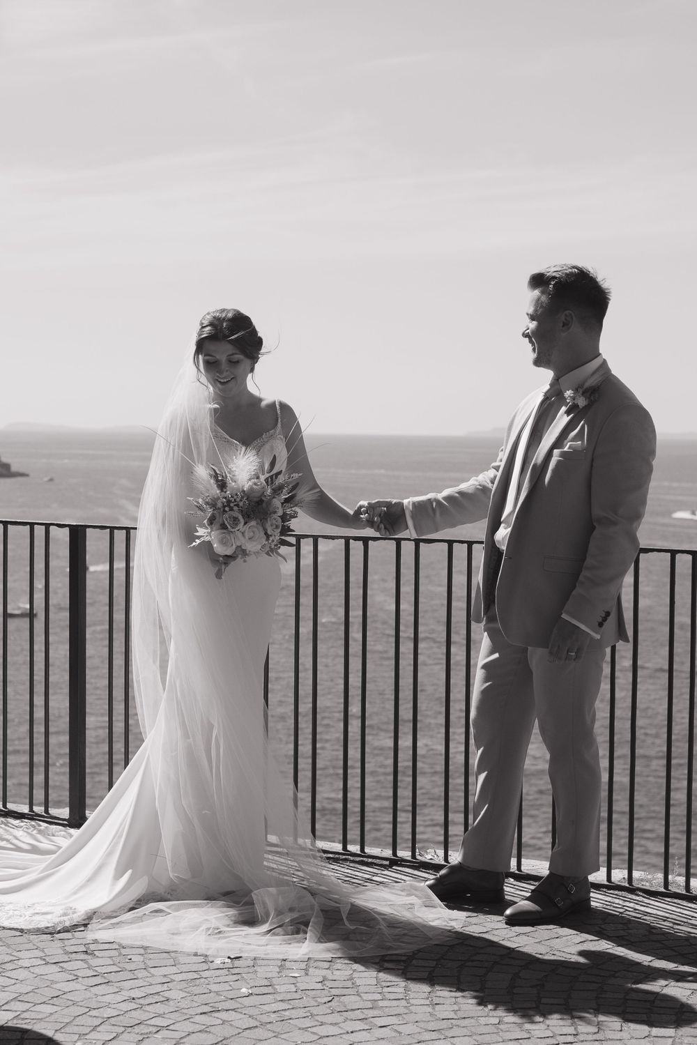 A black and white image of a newlywed couple on a balcony by the sea, holding hands. the groom is looking at the bride, bride looks down