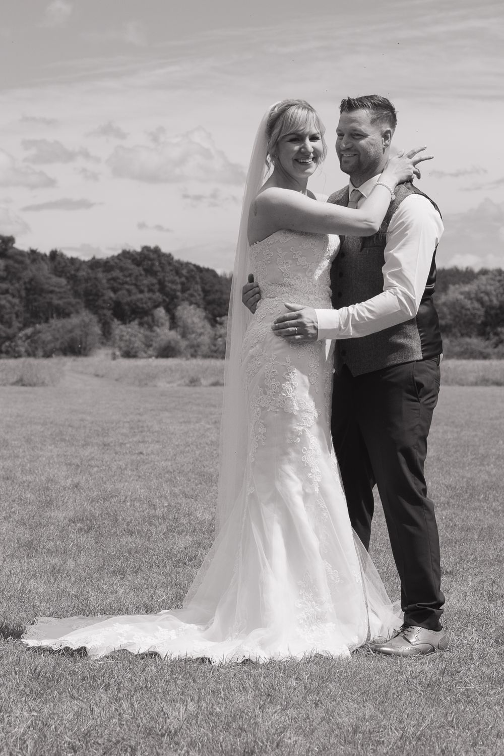 Black and white image of a bride and groom laughing as they have a cuddle, in an open field with trees in the background.