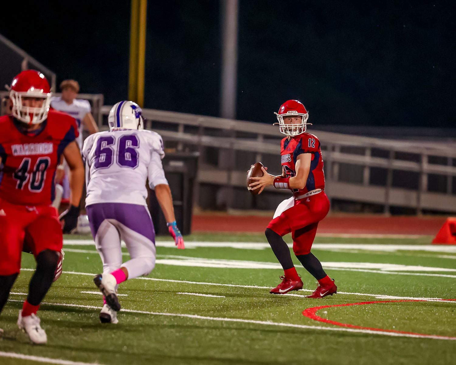 High school football game action shot showing offensive and defensive players on illuminated field at night.