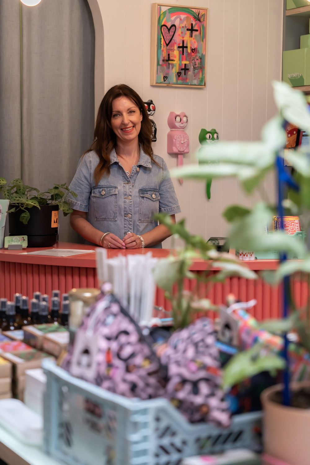 A small colorful retail shop interior with stocked shelves, pink decor and wall art, and houseplants.