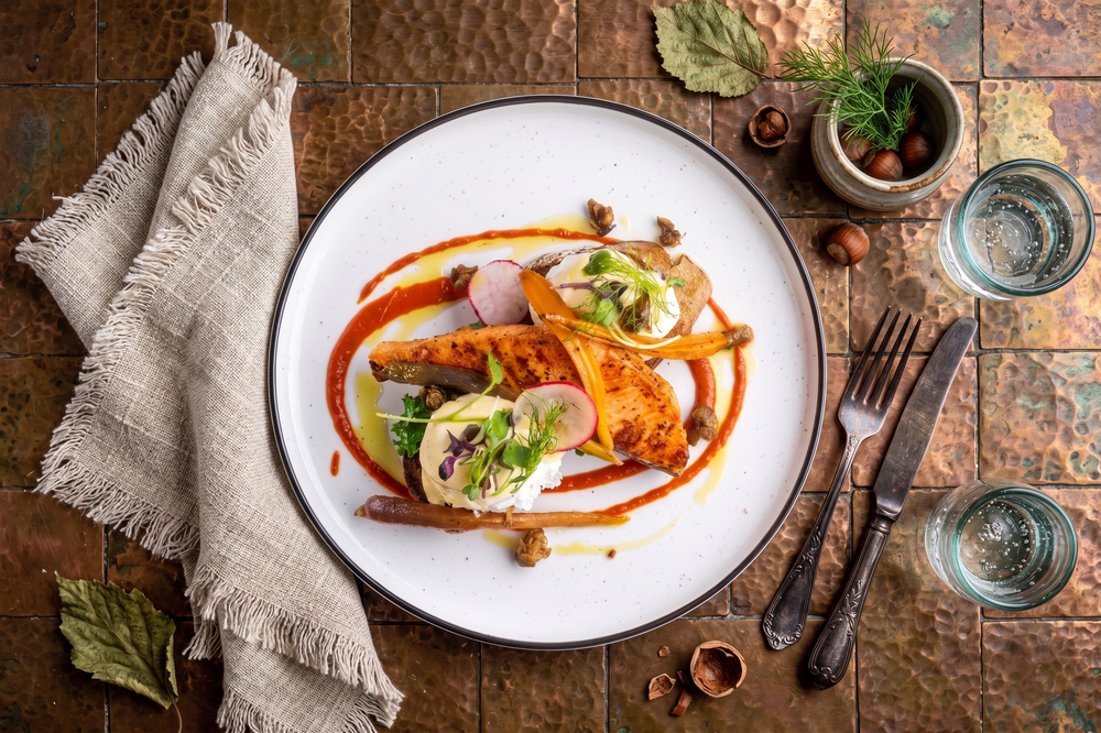 Gourmet dish with roasted vegetables, herbs, and sauce on a white plate, styled on a rustic copper surface.