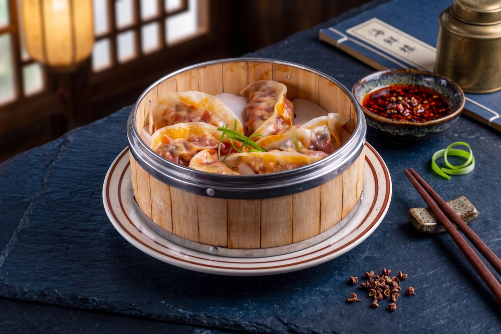 Steamed dumplings in a bamboo steamer basket served with chopsticks and chili sauce on a blue stone table.
