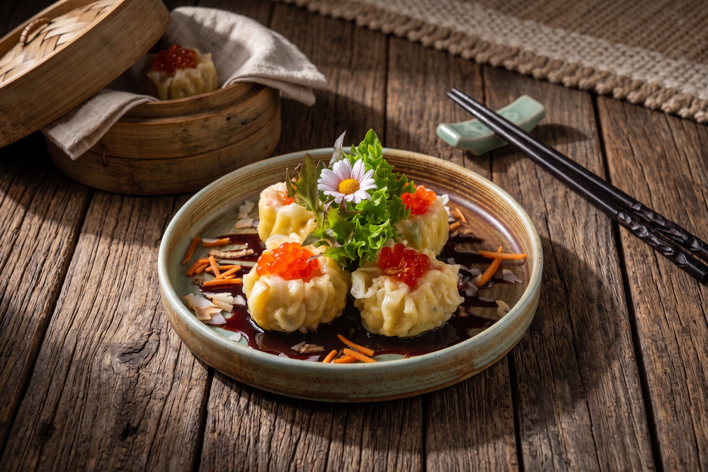 Steamed siu mai dumplings topped with fish roe, served in savory sauce with floral garnish on rustic wooden table.