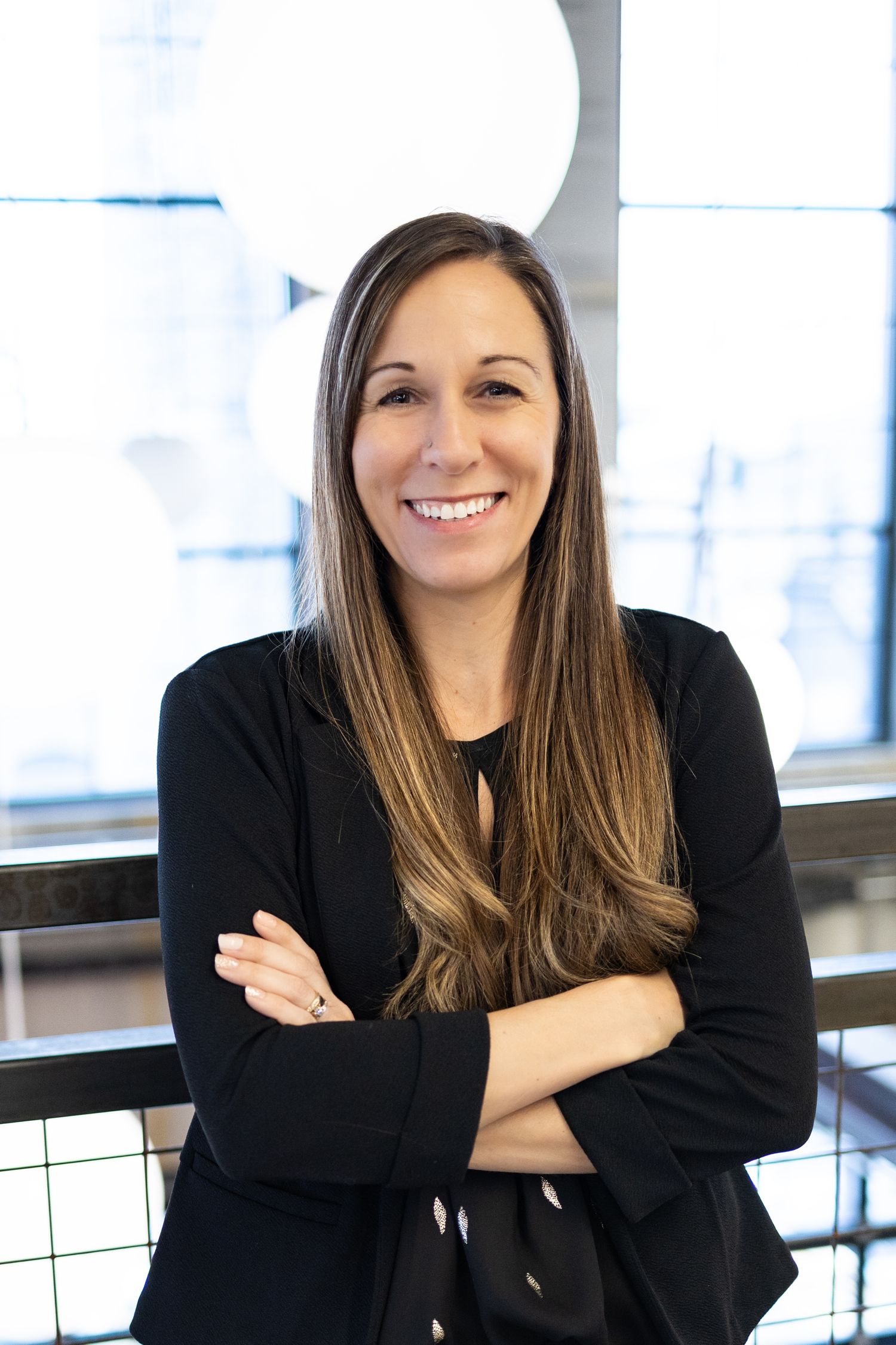 Professional portrait of woman in black blazer with long brown hair against bright office background.