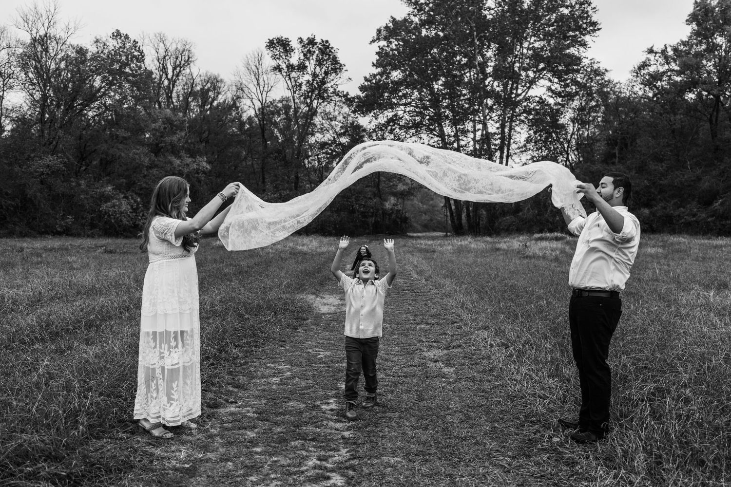 A child stands between two people holding a flowing white fabric in a black and white outdoor scene.