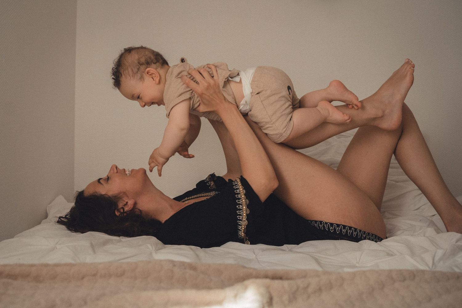 1 maman allongée sur son lit sourit à son bébé qu'elle tient au dessus d'elle et qui lui sourit aussi en tendant ses bras vers elle.Bonheur.