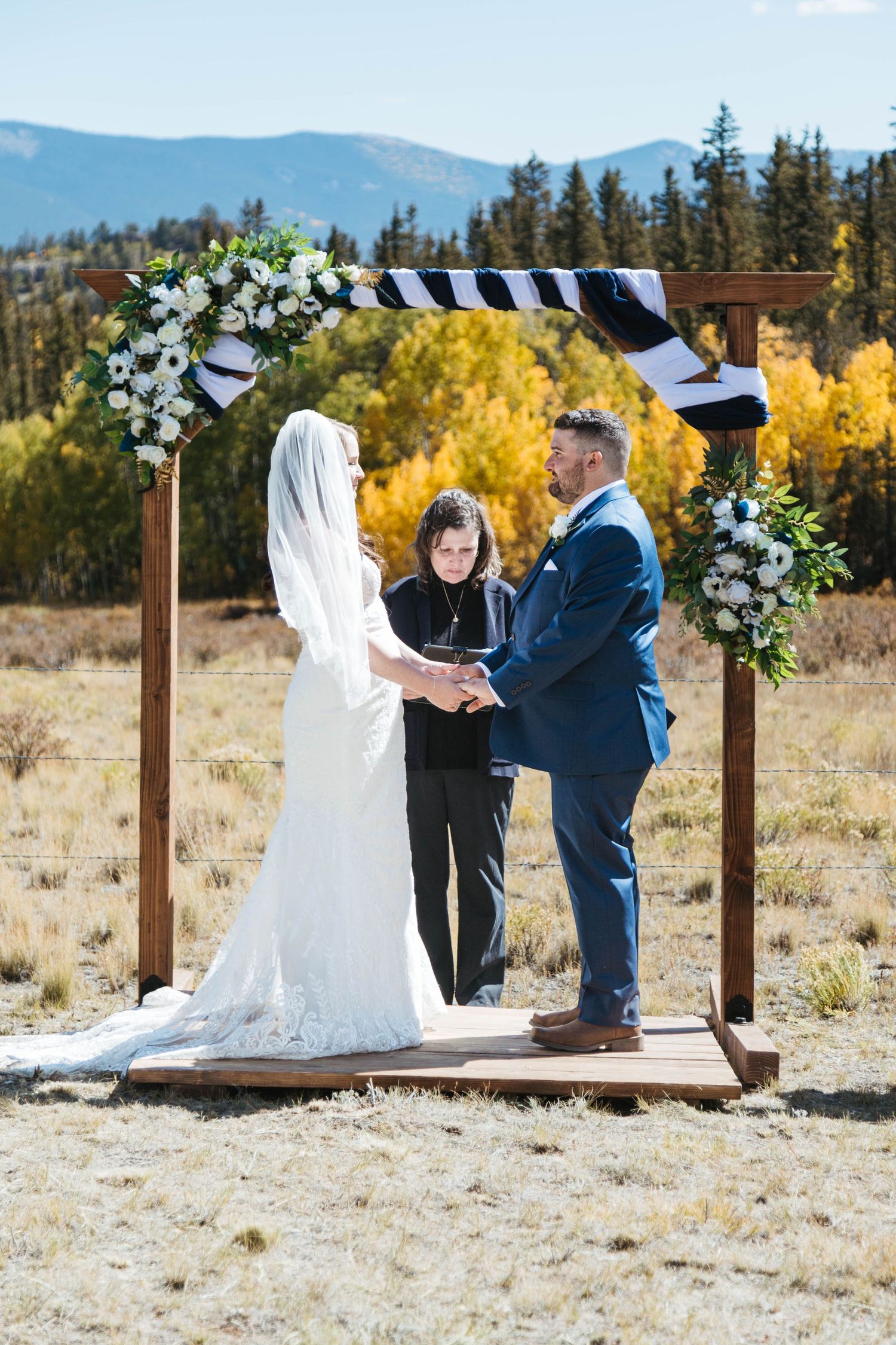 A wedding ceremony takes place under a wooden arch decorated with white flowers against a backdrop of autumn mountains.
