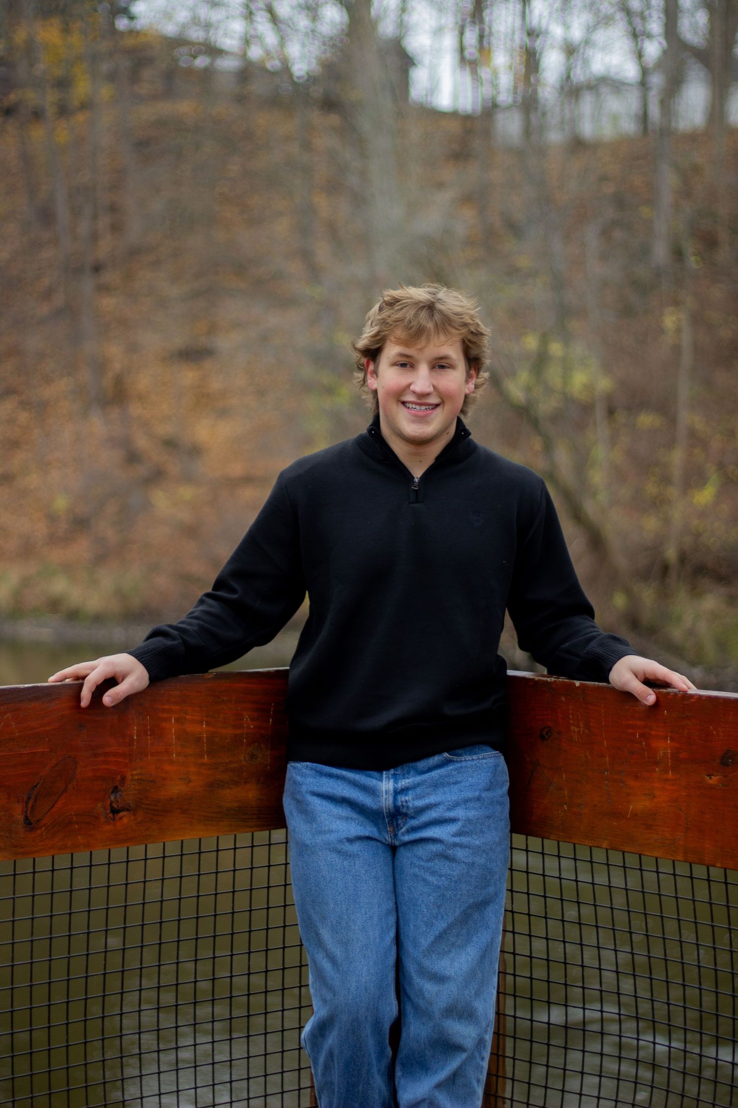 Someone in a black sweater and jeans leans against a wooden railing on an outdoor deck during autumn.