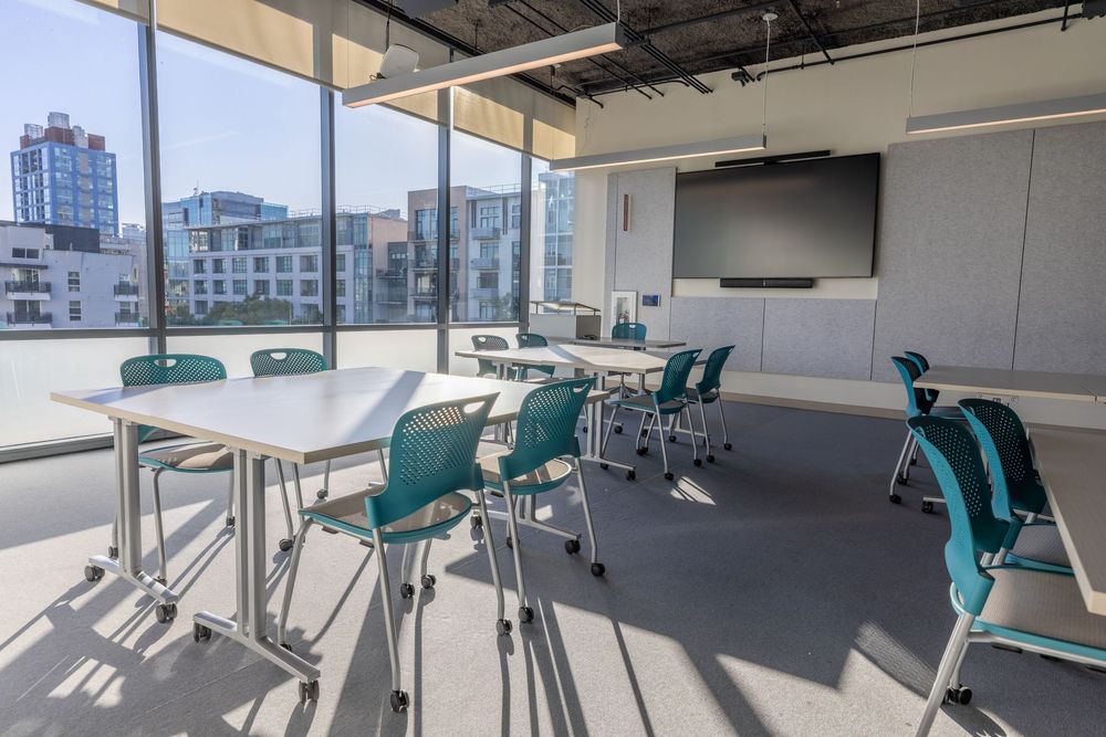 Modern office space with teal chairs and white desks illuminated by natural light through large windows.