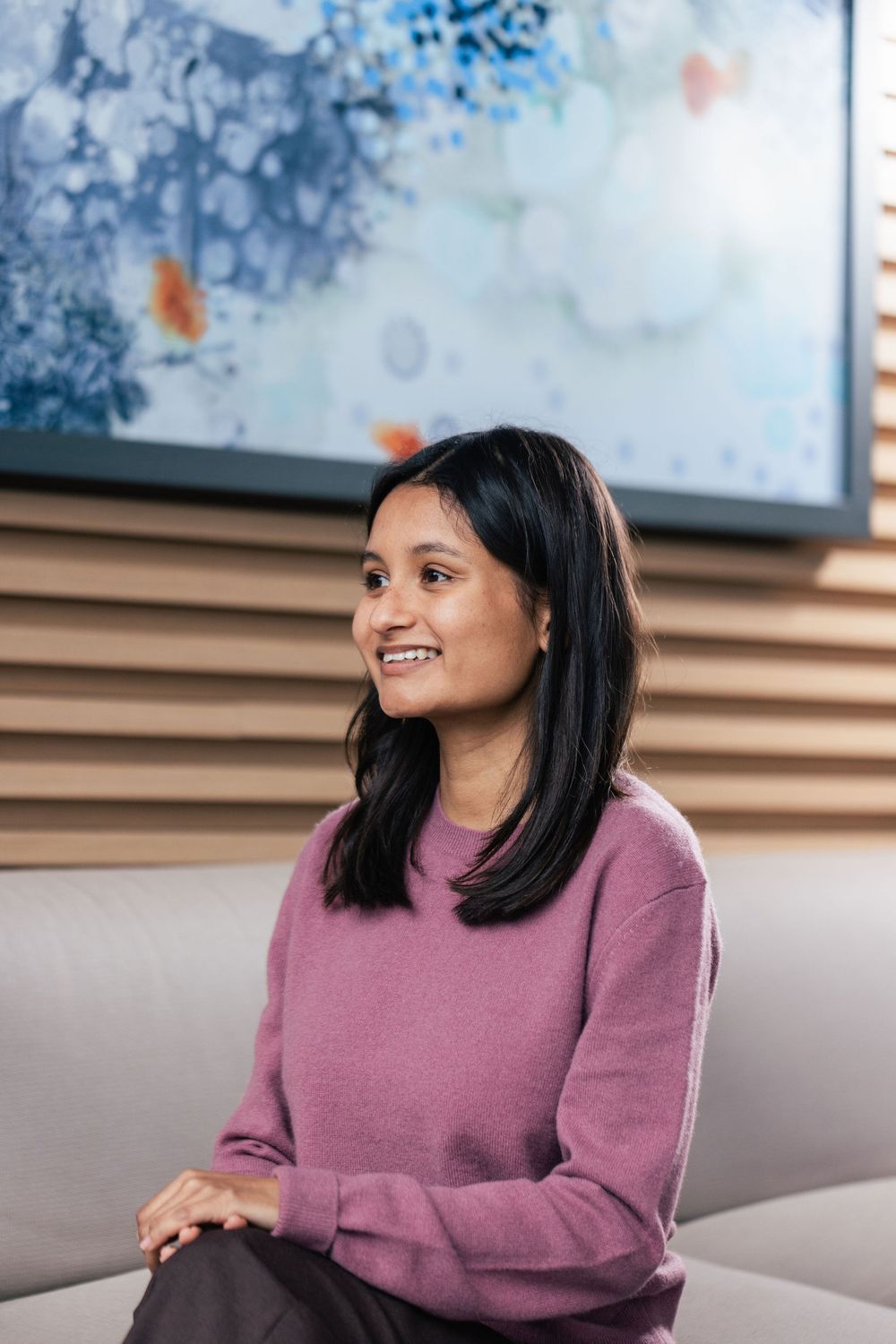 A person wearing a mauve sweater sits in an office space with abstract blue artwork on the wall behind them.