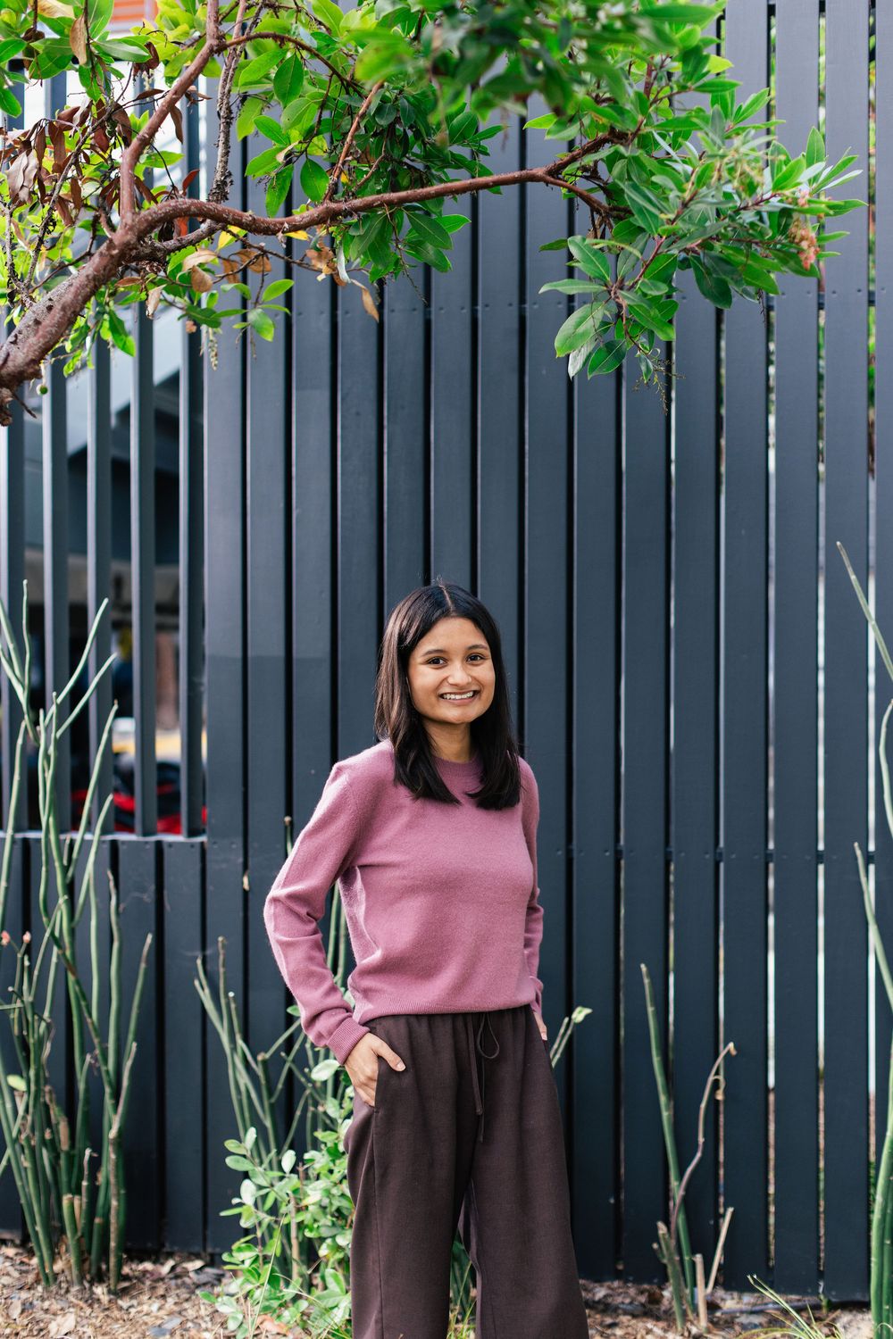 A person in a pink sweater and brown pants stands in front of a modern dark fence with green foliage.