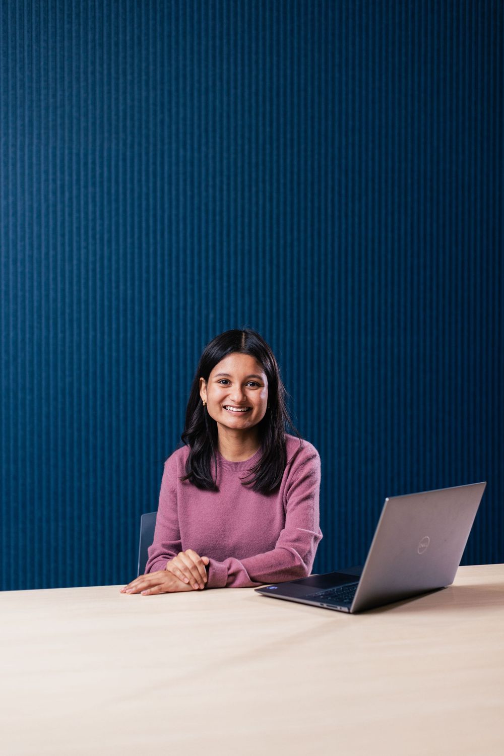 Person in mauve sweater sitting at desk with laptop against navy blue wall.